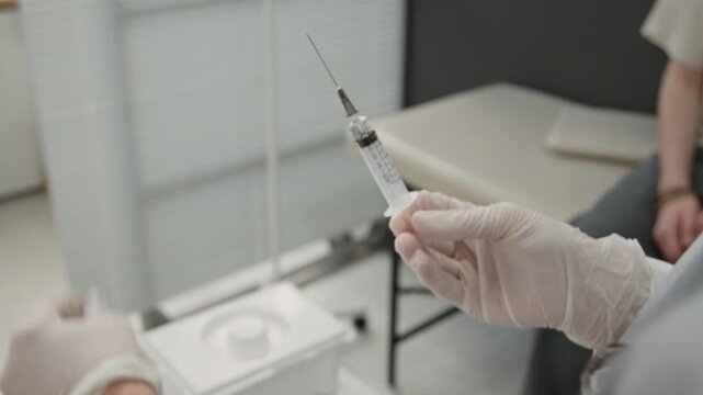 Close up of hands of mature Caucasian nurse wearing latex gloves preparing syringe with vaccine for young man sitting in background