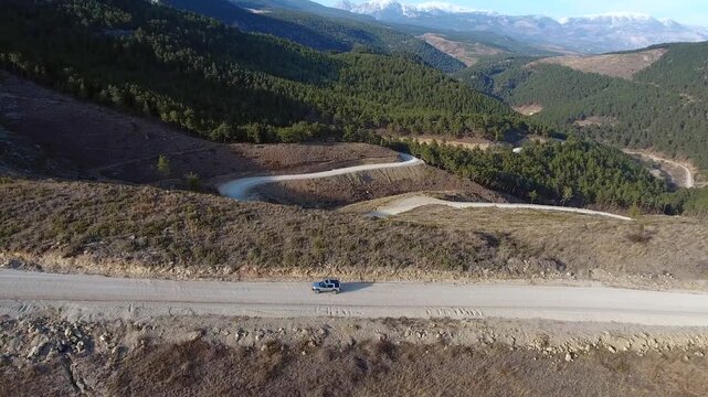 Aerial view of vehicle driving along flat mountain road toward dense green forest landscape. Remote travel scene shows lone car approaching woodland area across rugged highland terrain.