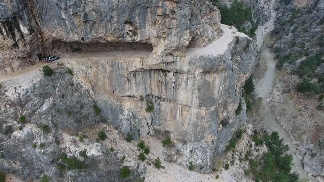 Aerial view of car driving along extremely dangerous rocky mountain cliff road track. Rugged vehicle follows a perilous ledge route above steep stone slopes in remote Himalaya.