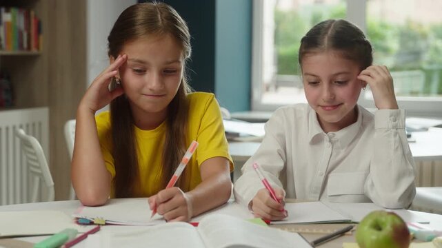 Concentrated two Caucasian little girls children solving difficult task together at desk inside class studying confused kids schoolgirls students pupils writing homework lesson education schoolwork