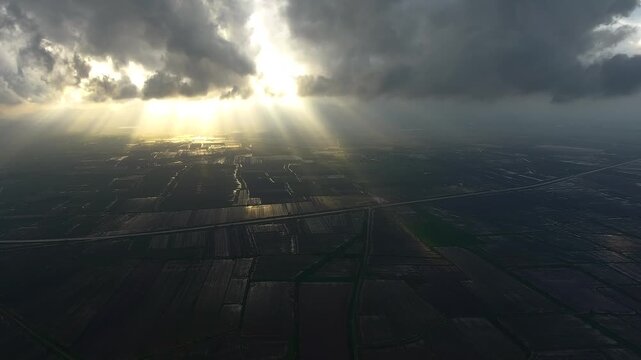 Aerial view of dark storm clouds casting dramatic light over flooded plains after rain. Sunbeams break through heavy cloudbank illuminating soaked cropland and wetlands.