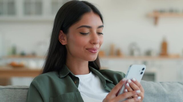 Happy surprised Indian woman using smartphone at home browsing social media saying wow at good news offer smiling amazed reaction Arabian female ethnic girl scrolling mobile phone chat online indoors