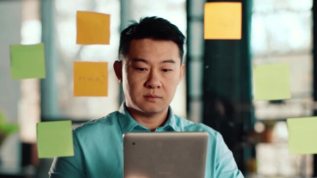 Man with dark skin stands in a bright office space, focusing on sticky notes attached to a glass wall. He uses a tablet to organize and plan ideas for a project.
