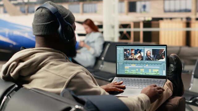 Digital nomad videographer on laptop in airport terminal lounge, cutting and editing video footage for web content while waiting for flight departure. Location independent nomadic lifestyle.