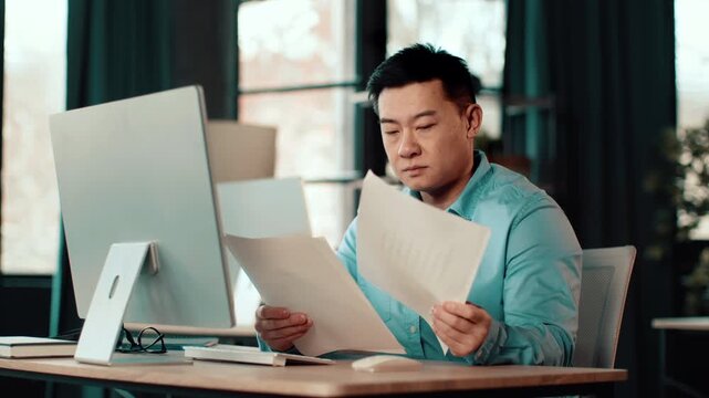 A man with light skin sits at a desk, looking closely at important documents in a modern office. The setting has large windows and a casual atmosphere with soft lighting.