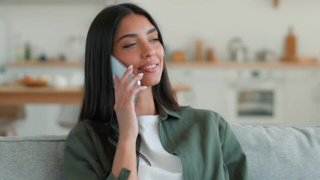 Happy laughing Muslim Arabian woman at home sofa talking on smartphone smiling having fun call friend Islamic female girl communicating relaxed positive mood using mobile phone conversation indoors