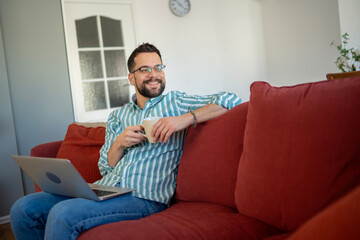 Obraz premium Smiling man relaxing on sofa, enjoying coffee and using laptop
