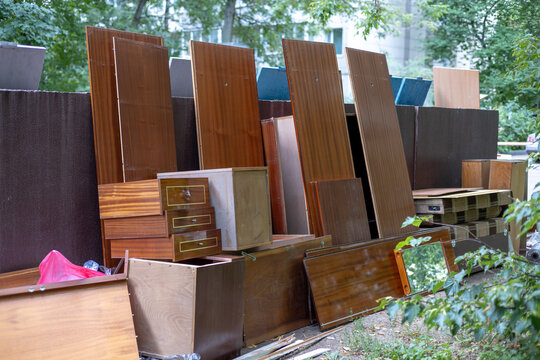 Dumped old outdoor furniture. Wooden cabinets, wooden doors, mirrors and shelves stacked against the wall of a city dumpster