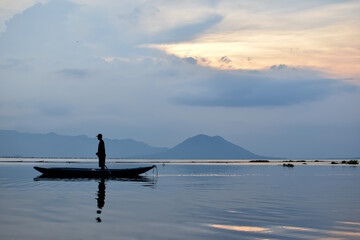 Fototapeta premium Minimalist scene of a fisherman on still water with mountains in the background.