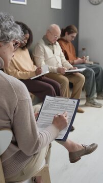 Vertical shot of female patient writing in medical paper form while waiting for health check up in queue in modern hospital, diverse people visible in background