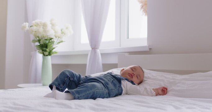Portrait of a sweet cute little baby wearing denim overalls sleeping at home lying on comfortable bed on white blanket. Adorable infant child napping alone resting in cozy bedroom. 4k video.