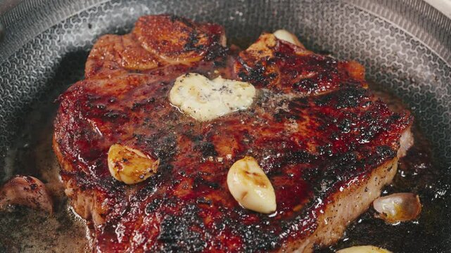 Close-up video of a T-bone steak sizzling in butter with garlic in a cast iron pan. Smoke rises as the steak cooks, highlighting rich texture, heat and a premium cooking experience, perfect for gourme