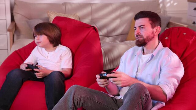 Emotional father and son with controllers playing video game on poufs at home
