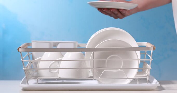 Woman putting clean dishes and cutlery into drying rack on white table against light blue background, closeup