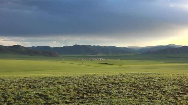 Mongolian ger village spreads across open steppe grassland beneath dark mountain ridge. Crowded yurt settlement lines the green plain, forming a nomad campsite near distant peaks.