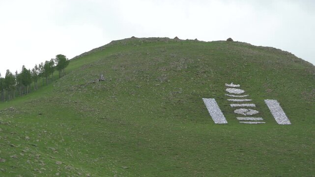 Mongolia mountain hillside marked by white stone Soyombo symbol, national emblem landscape. Mongol peak carries bright rock glyph on grassy slope, cultural sign seen from far away.