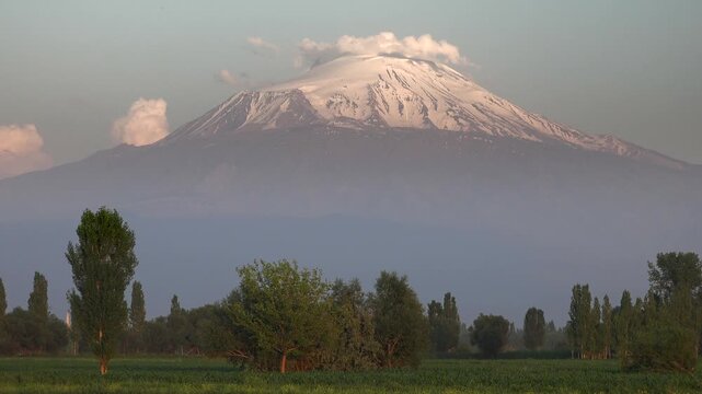 Time lapse shows the snowy peak of Mount Ararat rising beyond a wide green plain at dawn. Cloud movement and distant summit light define the monumental landscape in eastern Turkey.