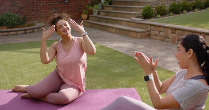 Clapping woman in gray mirroring gestures beside pink-clad woman on backyard lawn, purple mat