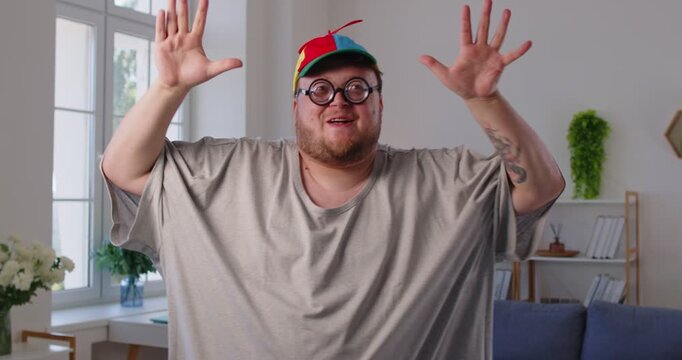 Chubby overweight man dancing goofy at home livingroom. With glasses and a propeller cap, he shows funny, silly moves having fun in apartment, radiating happy vibes and body positivity. 