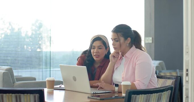 Diverse female coworkers leaning, pointing and typing at laptop, opening brief and collaborating