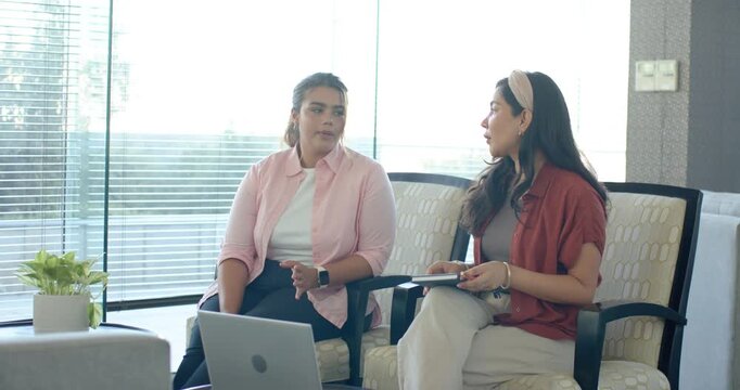 Female coworkers talking about work in office right holding tablet explaining left glancing laptop