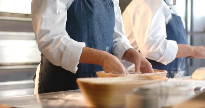 Diverse female bakers placing dough into baskets for proofing upon bowl arrival at bench in aprons