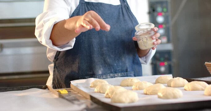 Mid adult baker wearing apron sprinkling flour from jar over rolls at prep counter before baking