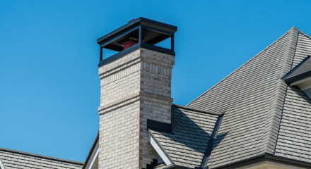 Obraz premium Brick chimney with metal cap on residential house roof against a clear blue sky in daylight; architectural detail of roofing structure