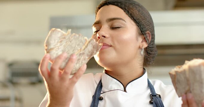 Woman baker in chef coat holding two loaves lifting left smelling crust assessing aroma in bakery