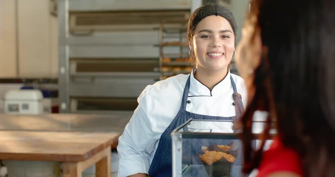 Diverse female baker in chef coat turning to customer entering, ordering cupcakes at glass case