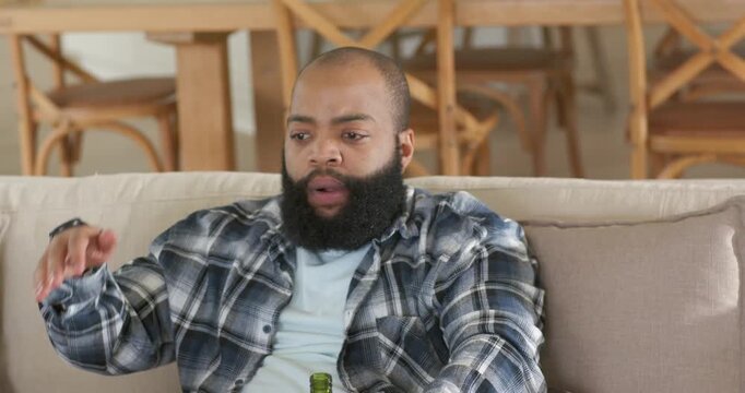 African American man sitting on couch holding beer bottle leaning on soccer ball rubbing forehead