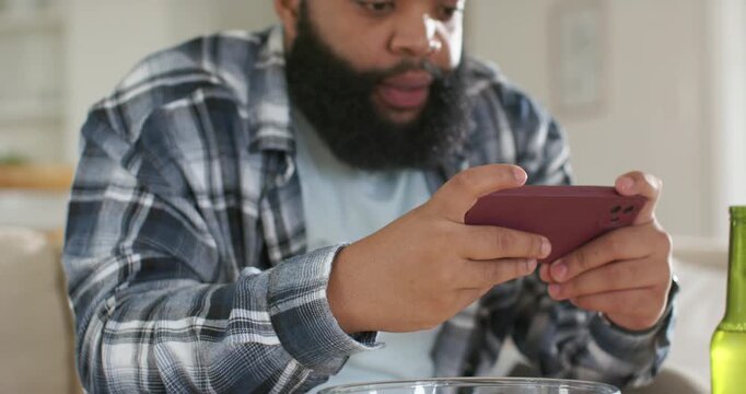 Bearded man tapping maroon phone and eating popcorn on couch, animated UI showing clips for media