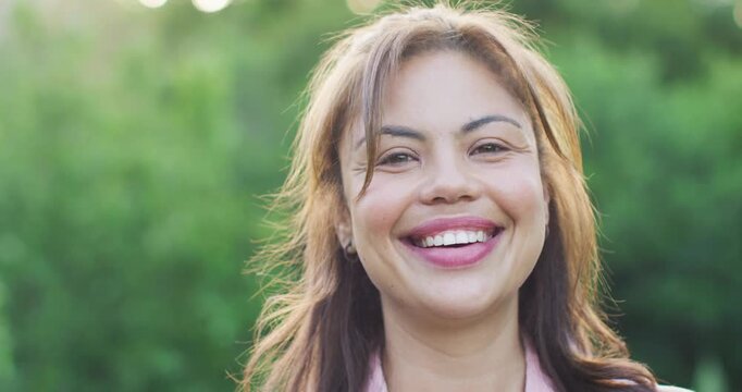 Smiling woman tilting right, blinking and keeping gaze in park, in light-pink blouse and studs