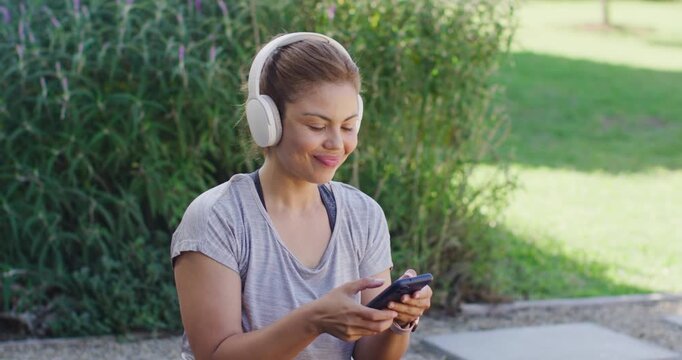Lowering smartphone, woman meditating on park step, breathing, wearing tee, headphones, tracker