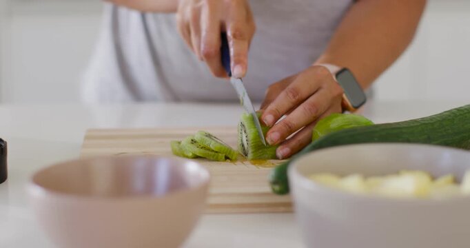 Woman steadying halved kiwi, slicing thin rounds with paring knife on wood board making snack