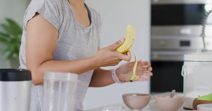 Woman is inspecting banana at kitchen counter, is pulling peel downward in sections for blender cup