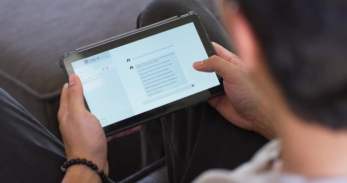 Asian man sitting on sofa positioning tablet on knees and scrolling messaging thread with thumb