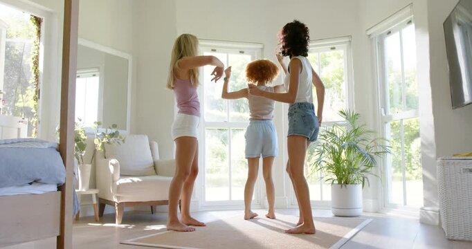Three adult female friends rehearsing dance on cue in sunlit room by bay windows wearing shorts