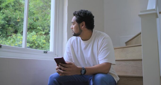Man sitting on stairs in white tee, jeans using phone, reacting, looking out window, smiling