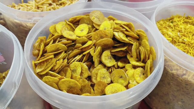 Camera orbiting around a container of crispy banana chips, showing golden fried slices coated with spices alongside other Indian snack mixtures in a market display!