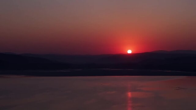 Cinematic fixed aerial shot of a vibrant red sun setting behind mountains with a bright reflection on a calm river in China.