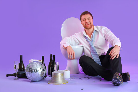 Drunk young businessman sitting near toilet bowl after party on lilac background