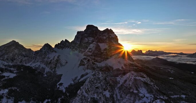 Sun rises behind silhouetted mountain peak in winter landscape with drone panning over snowy terrain.