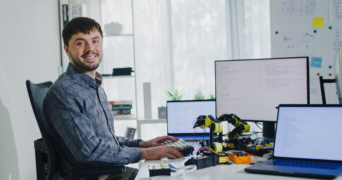 Portrait confident smiling male programmer working with robotics code AI software and looking at camera in tech office. Artificial intelligence dashboard, robotics, user interface, machine learning.