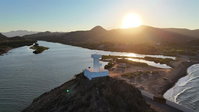 4K drone wrapping around a coastal lighthouse over an estuary inlet in Muleg&eacute;, revealing a natural sand and rock access with rich sunset colors.