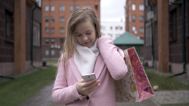 Handsome smiling young blonde girl in pink panto with mobile phone outdoors, with packages after shopping, have fun and enjoy