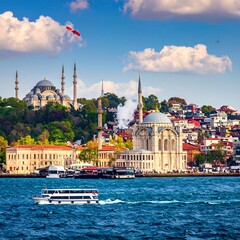 Naklejka premium Scenic waterfront view showcasing mosques, buildings, watercraft under a blue, cloudy sky