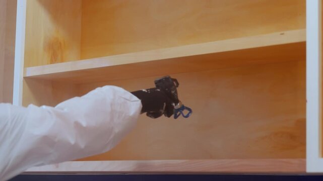 Worker using a spray gun to apply paint or coating inside cabinet during renovation. Close up of professional finishing process
