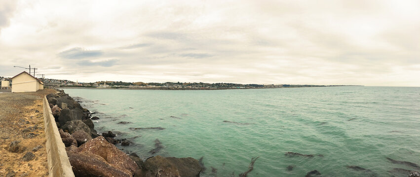 Panoramic coastal view of Friendly Bay in Oamaru with turquoise waters, rocky breakwater, and South Hill cityscape under cloudy sky - Otago, New Zealand, February 5, 2017