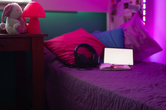 Teenager bedroom showing modern aesthetic with purple neon lighting, tablet, headphones on a bed and cozy pillows inside a contemporary interior styled for youth